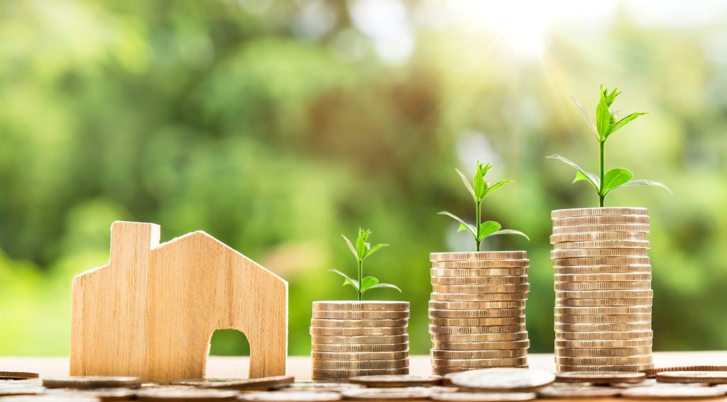 a small wood block house next to stacks of coins with tiny trees on top